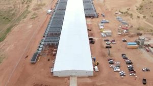 An aerial view of a massive, long white data center building surrounded by rows of cooling infrastructure in the dusty red dirt of Lubbock, Texas.
