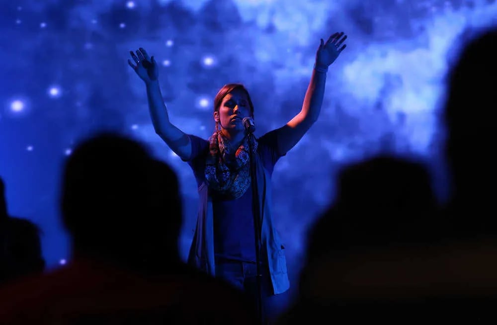 A woman stands on a stage with her arms raised during a worship service at a modern megachurch, illuminated by deep blue stage lighting against a starry digital backdrop, with the silhouettes of a congregation in the foreground.