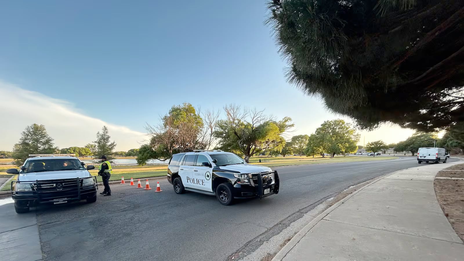 A Lubbock Police Department SUV and a public service vehicle blocking off a road next to a grassy park area with orange traffic cones.