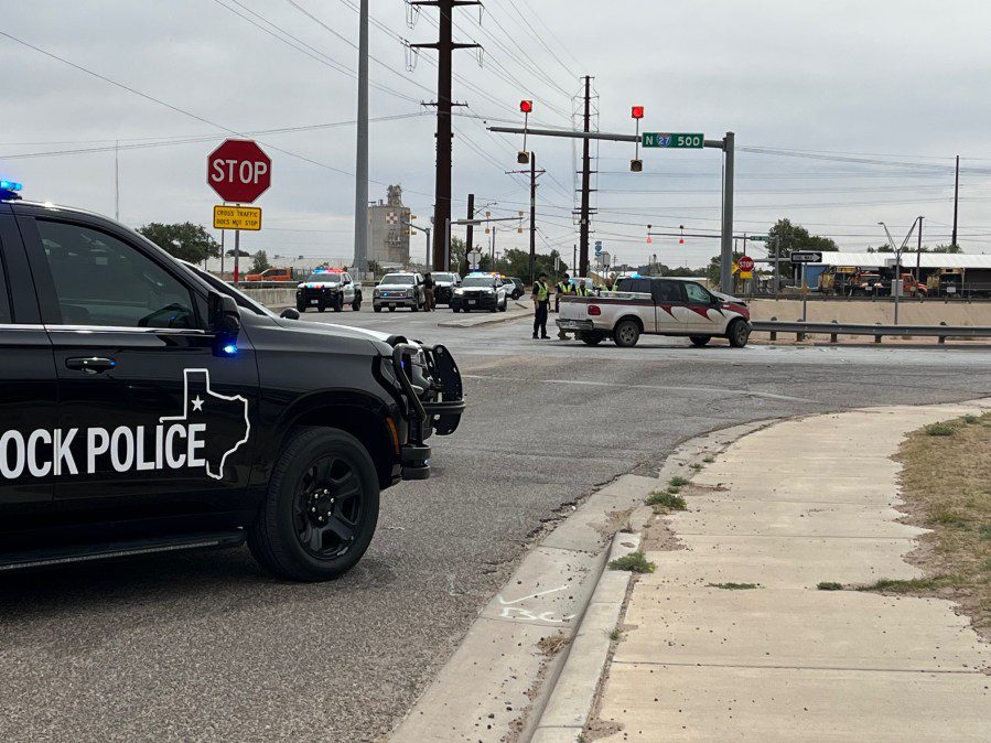A Lubbock Police Department vehicle with flashing blue lights parked at an intersection next to a damaged white pickup truck under the I-27 overpass signs.