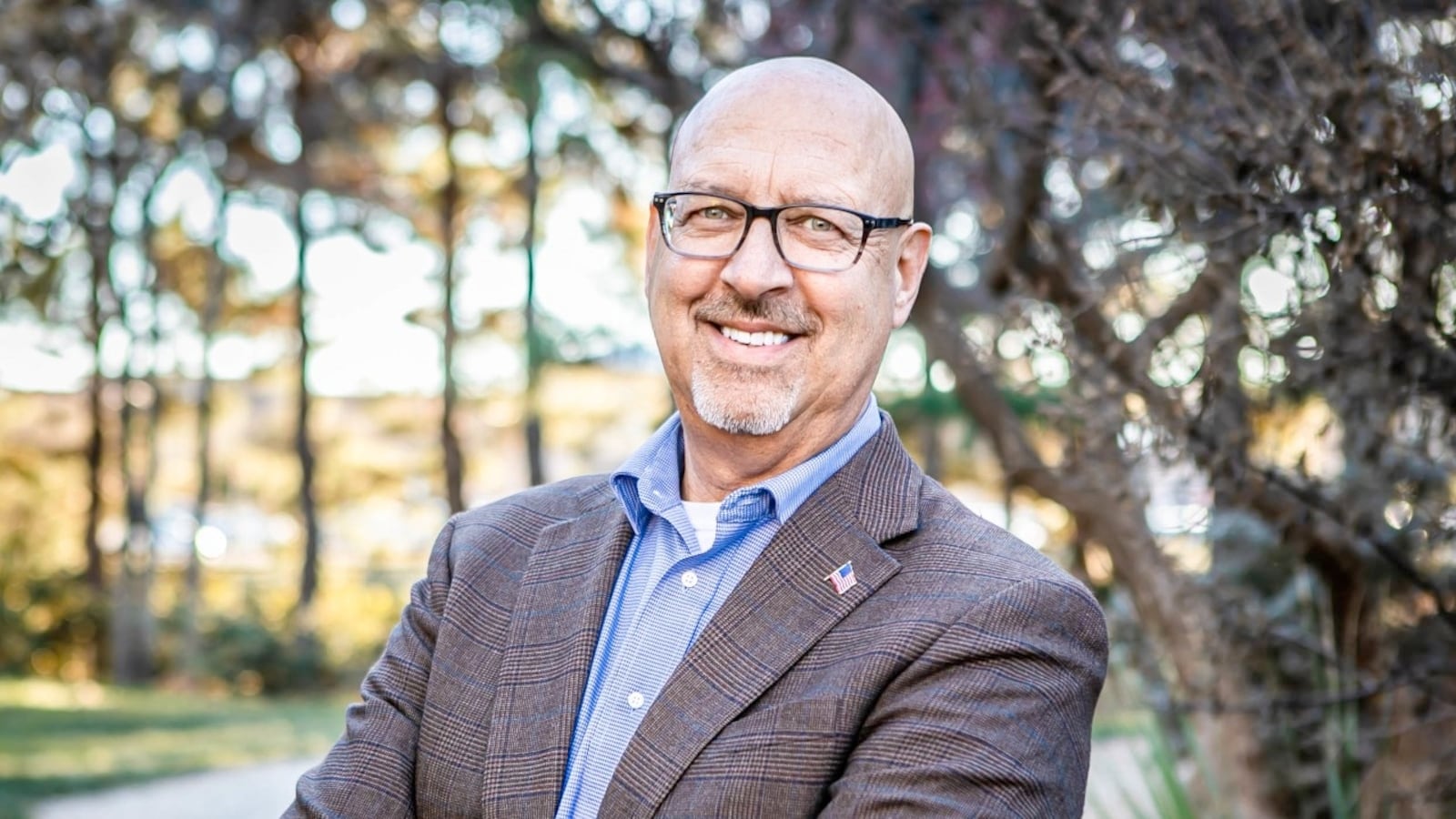 Mark McBrayer, the Mayor of Lubbock, smiling with the radiant confidence of a man who knows most of his constituents forgot it was Election Day. He is wearing a patterned blazer and a tiny American flag pin, standing in front of some blurry trees that are likely the only green things left in town after the latest dust storm.