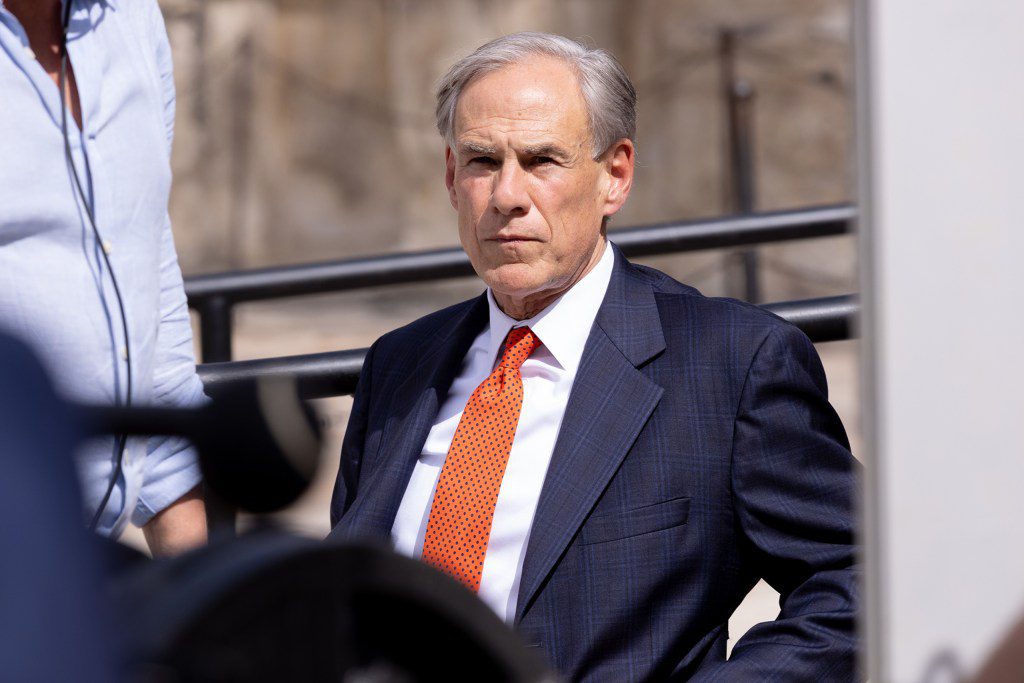 Texas Governor Greg Abbott wearing a blue plaid suit and a bright orange tie, looking intensely serious.