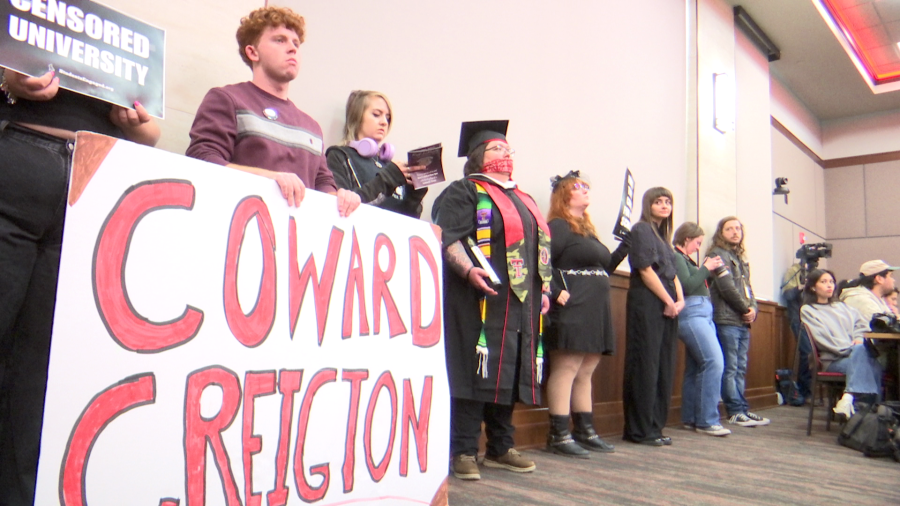 Students and faculty at a Texas Tech Board of Regents meeting holding a "Coward Creighton" sign while a student in a graduation gown wears a red gag over their mouth.
