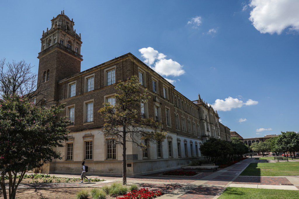 The exterior of the brick Texas Tech University Administration building with its prominent tower under a blue sky, as a student walks along the campus pathway.