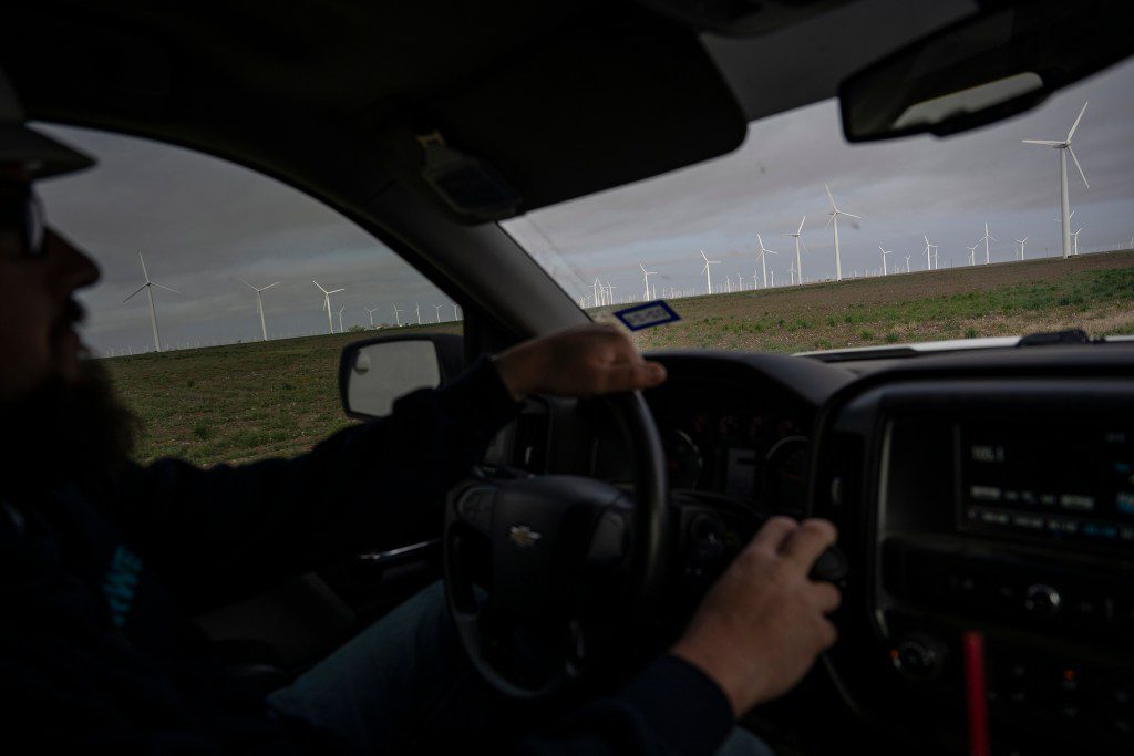 A view from inside a Chevy truck driving past a massive field of wind turbines under an overcast West Texas sky.
