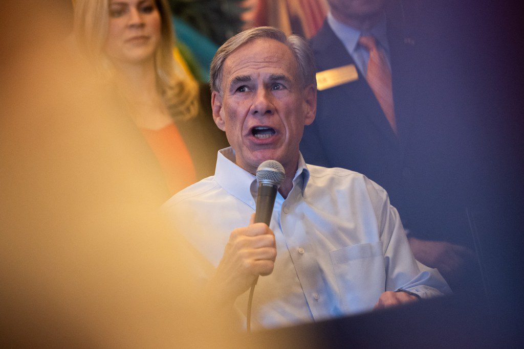Governor Greg Abbott speaking intensely into a microphone at a campaign event, surrounded by onlookers.
