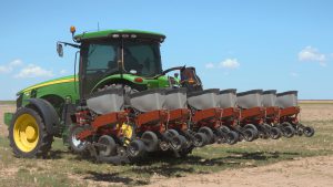 A green and yellow John Deere tractor equipped with a multi-row seed planter attachment parked in a dry, dusty agricultural field under a clear blue sky in West Texas.