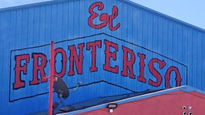 A blue corrugated metal building featuring a large hand-painted red sign that reads "El Fronteriso" with a red pole and satellite dish in the foreground under a clear sky.