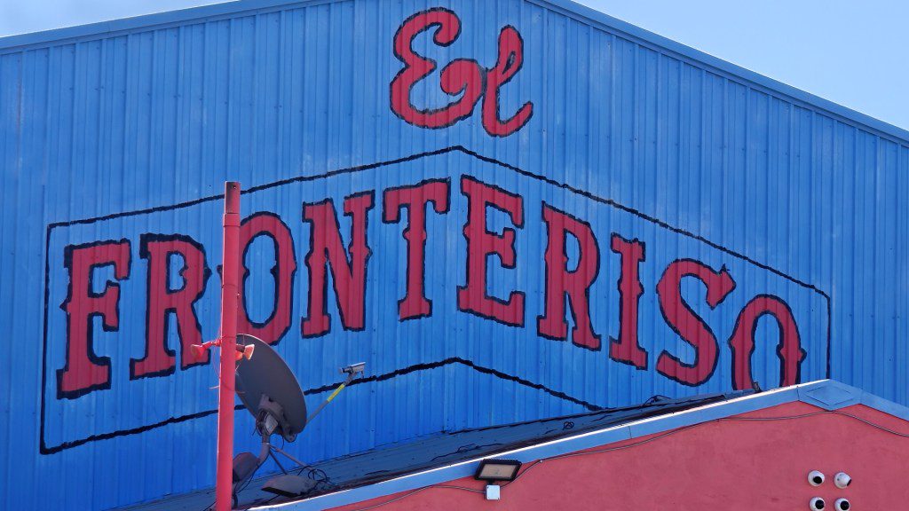 A blue corrugated metal building featuring a large hand-painted red sign that reads "El Fronteriso" with a red pole and satellite dish in the foreground under a clear sky.