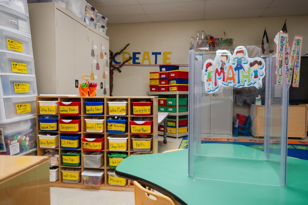An organized early-childhood classroom in Texas featuring colorful plastic storage bins, a green activity table with a plastic safety divider, and a decorative sign that reads "CREATE" on the back wall.