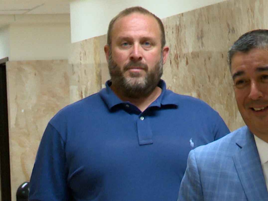 Joshua Allen, wearing a blue polo shirt, walking down a marble courthouse hallway with a slight smirk alongside another man in a plaid suit.