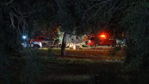 Emergency rescue vehicles with flashing red lights parked near a dark, wooded area at night where a dive team is recovering a vehicle from floodwaters.