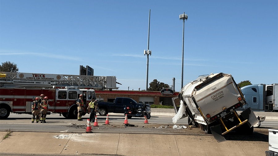 A City of Lubbock garbage truck rolled over on its side next to an access ramp on South Loop 289, with emergency crews on scene in front of a Crash Champions sign.
