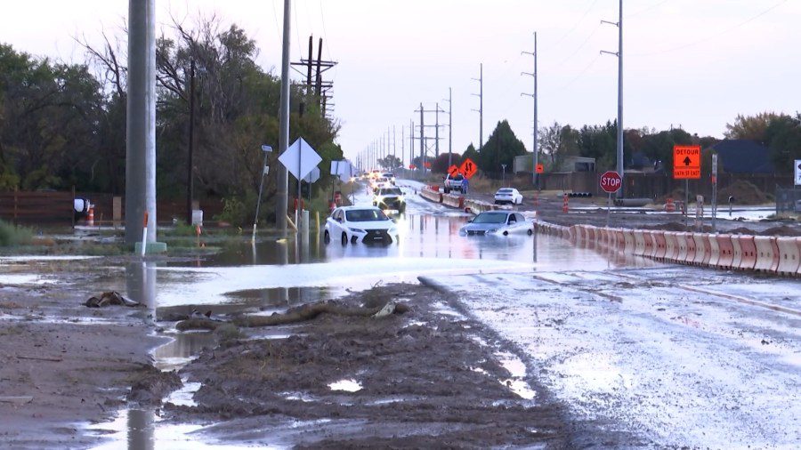 Two cars stranded in deep floodwaters on a Lubbock road surrounded by orange construction barriers.