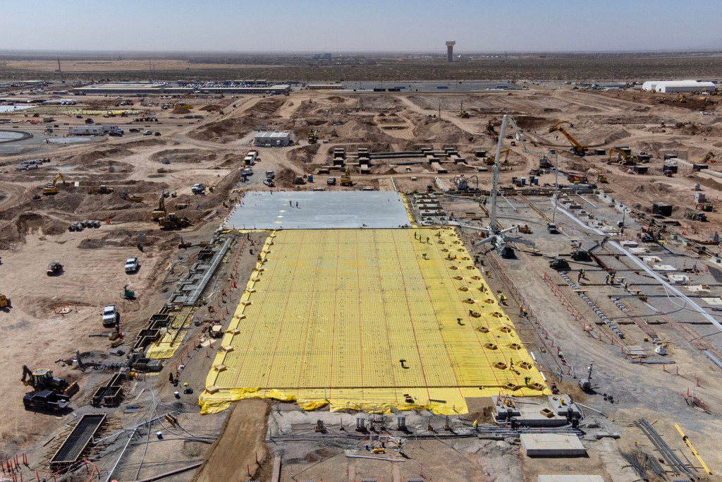An aerial view of the Meta Platforms data center complex under construction on a massive 1,000-acre site in far Northeast El Paso, near the Texas-New Mexico state line. The image shows heavy machinery, cranes, concrete foundations, and a large grid section covered in yellow vapor barrier sheets, captured on March 12, 2026.