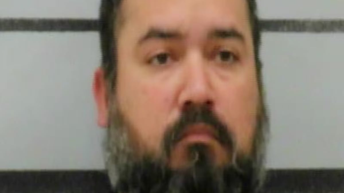 Mugshot of Pedro Ortiz with a dark beard and mustache looking directly at the camera against a gray striped background.