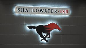 A backlit Shallowater ISD sign featuring a black and red running mustang logo mounted on a grey cinderblock wall.