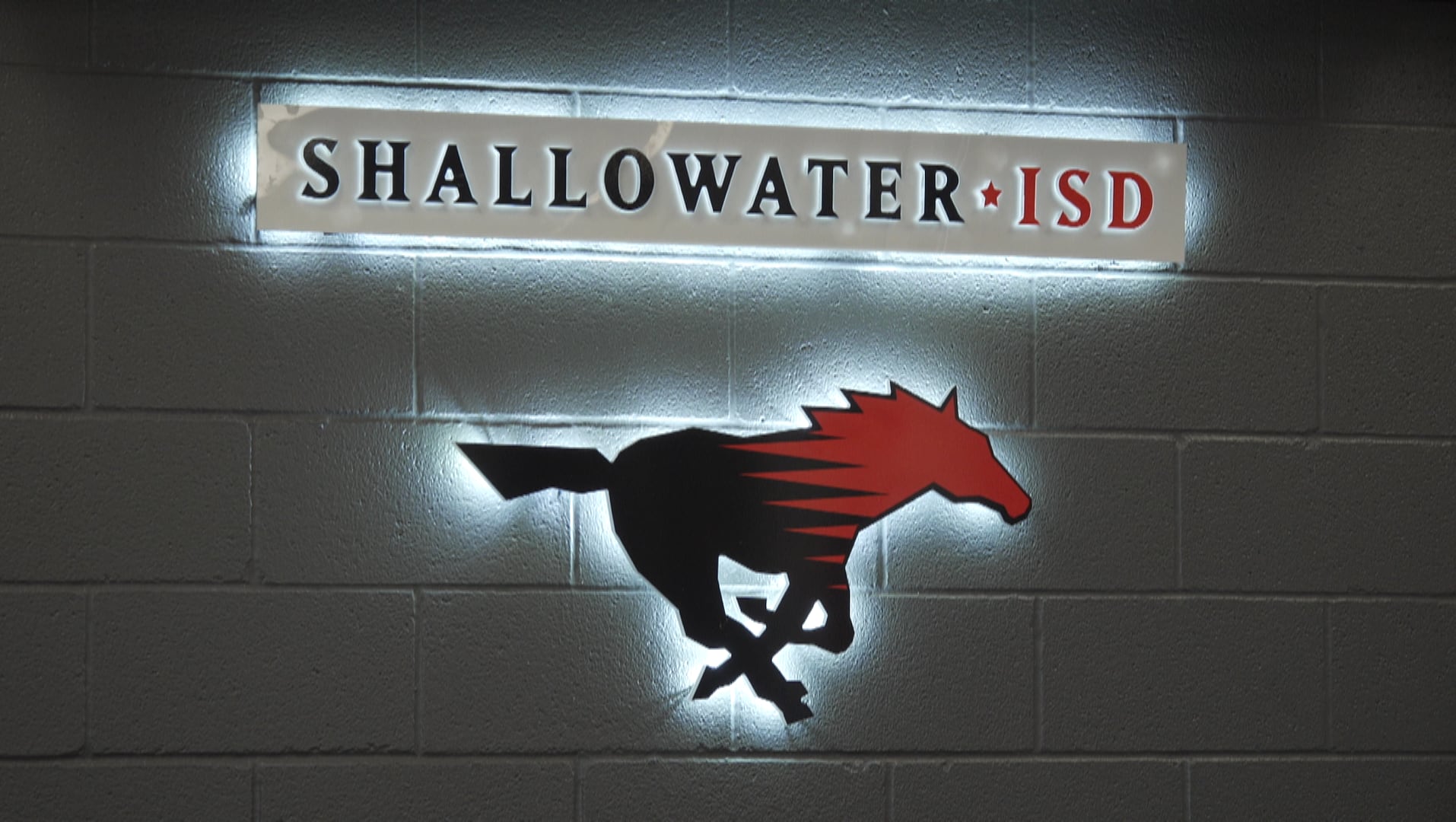 A backlit Shallowater ISD sign featuring a black and red running mustang logo mounted on a grey cinderblock wall.