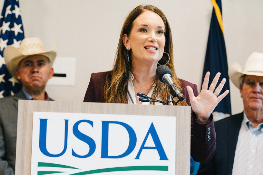 SDA Secretary Brooke Rollins speaking at a podium with a USDA logo during a press conference about the New World screwworm outbreak, flanked by men in cowboy hats.