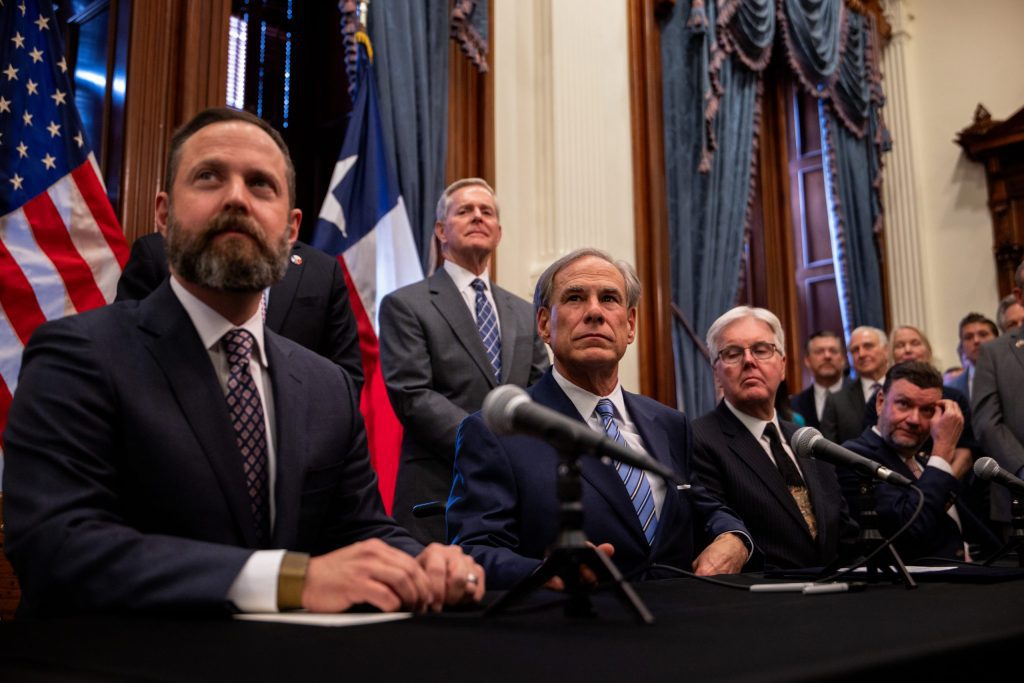 Texas House Speaker Dustin Burrows, Governor Greg Abbott, and Lieutenant Governor Dan Patrick sitting at a press conference table looking serious during a bill-signing ceremony.