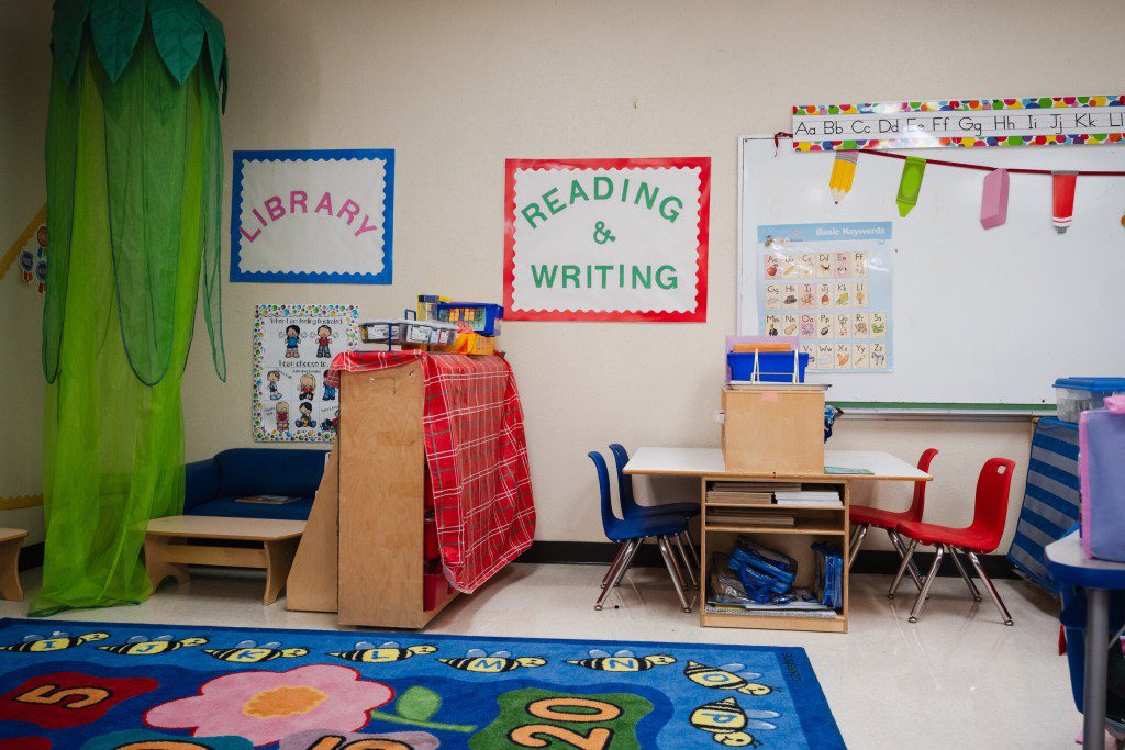 A colorful elementary school classroom with small red and blue chairs, a reading and writing sign on the wall, and a learning alphabet board.