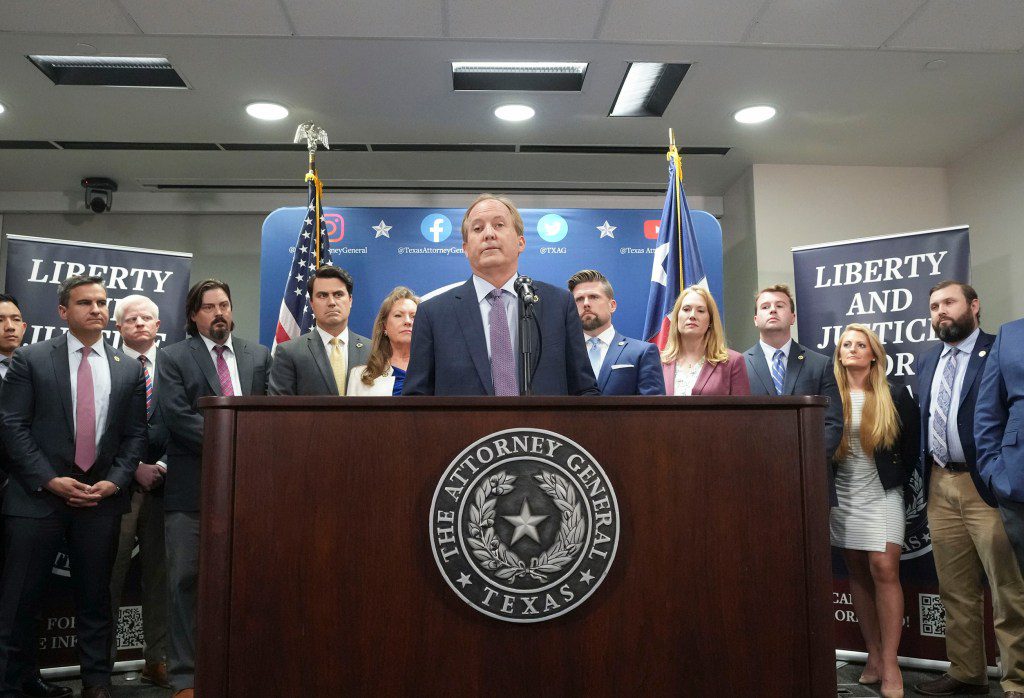 Texas Attorney General Ken Paxton speaks at a podium emblazoned with the state seal, surrounded by members of his legal staff during a press conference.
