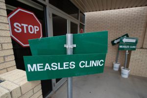 A green directional sign with white lettering that reads "MEASLES CLINIC" stands outside a brick facility, with a large red stop sign visible in the window of a glass door entryway behind it.