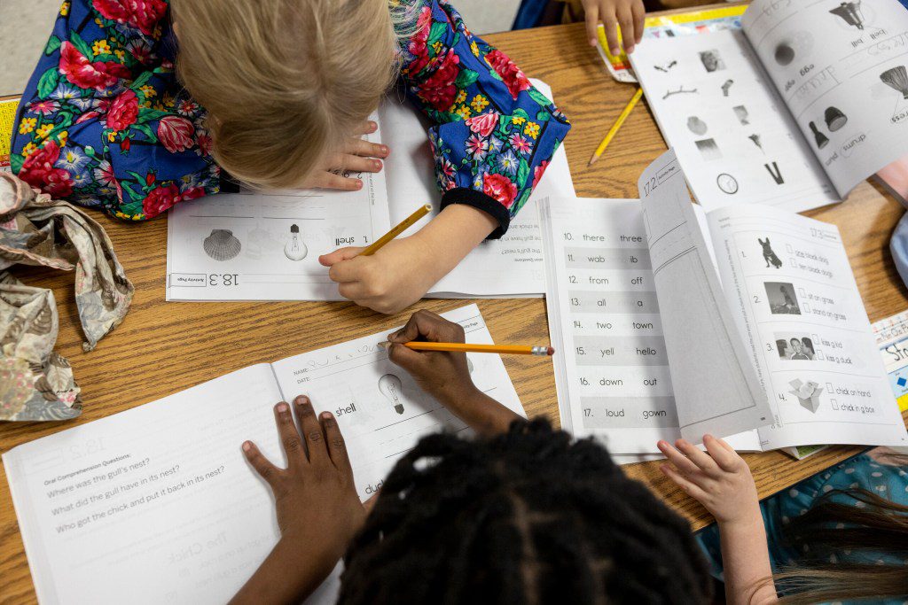 Elementary school students working on reading and writing assignments at wooden classroom desks.